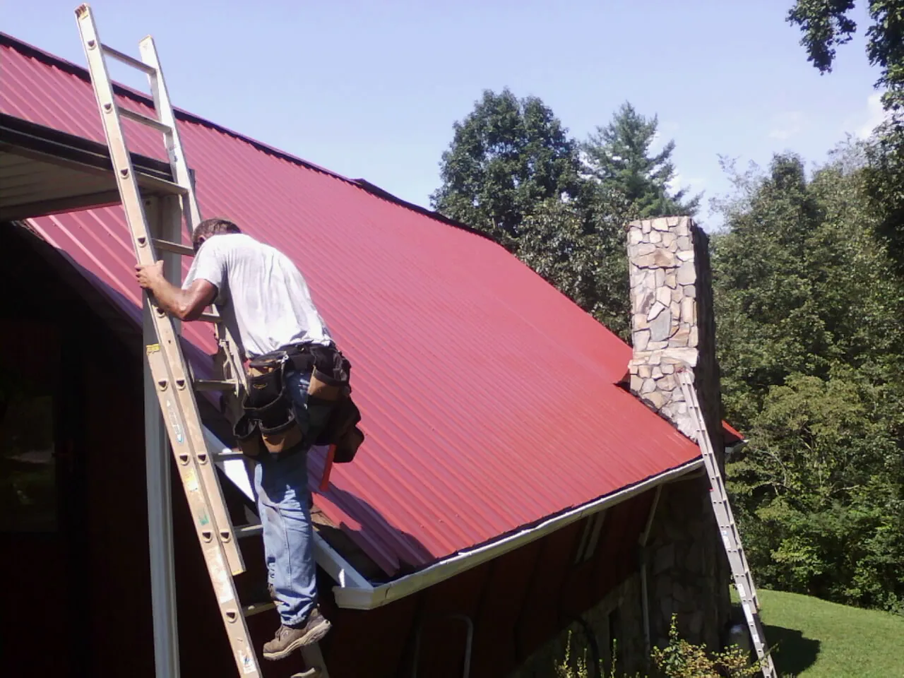 Roofer on a ladder beside a red metal roof and stone chimney — Soffit & Fascia Repair by Roofing Brothers in Nanjemoy